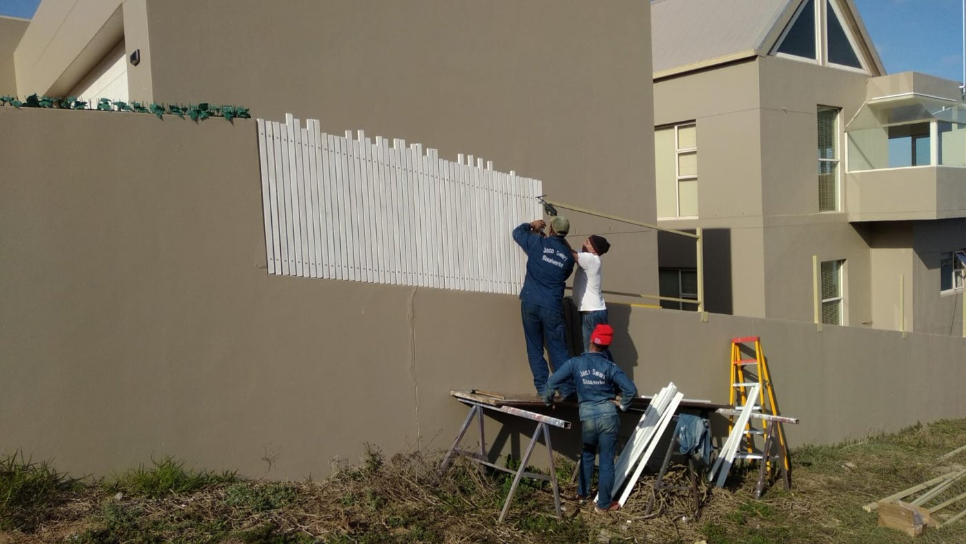 Three workers in Jaco Swart Staalwerke overalls fitting white slats to a boundary wall on site